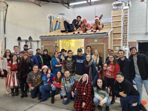Team Holiday Photo in warehouse three guys on a miniature house with candy cane 30 other team members standing in front of house