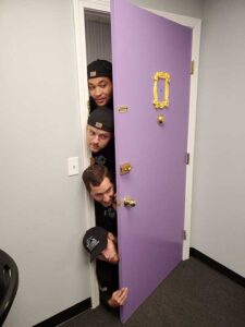 Team Photo 4 guys peaking vertically into a purple door
